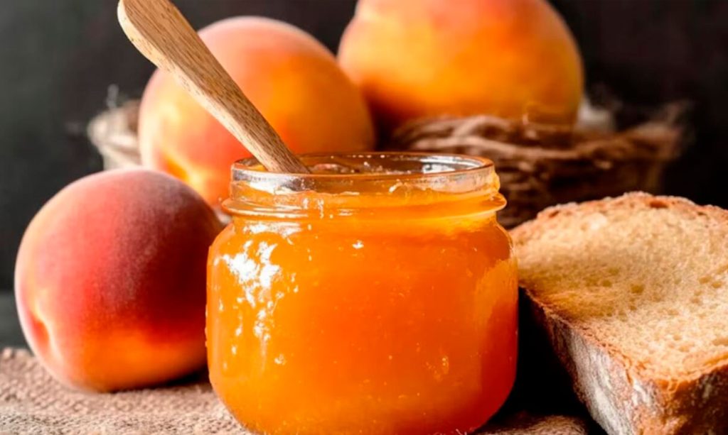 Tarro de mermelada de durazno casera D'La Abuela con duraznos frescos y pan tostado al fondo, sobre un paño de lino.